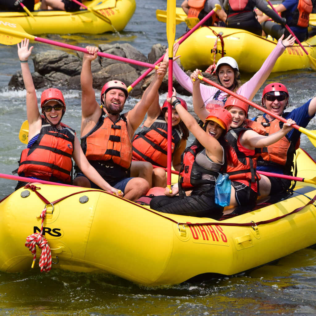 A group of people joyfully paddling in a bright yellow raft on a river, with arms raised and wearing orange life jackets.