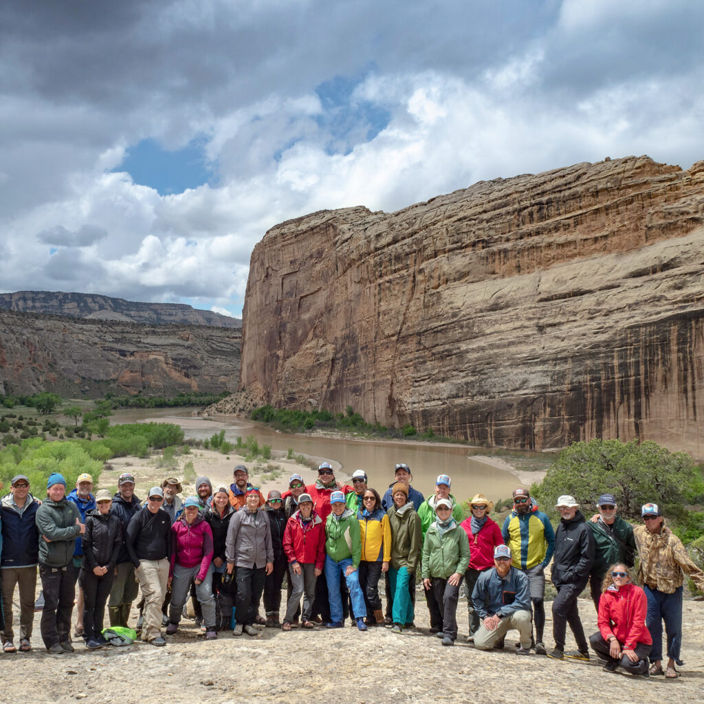 A group of people poses together in front of towering rock formations with the Yampa River in the background, under a cloudy sky in a scenic landscape.