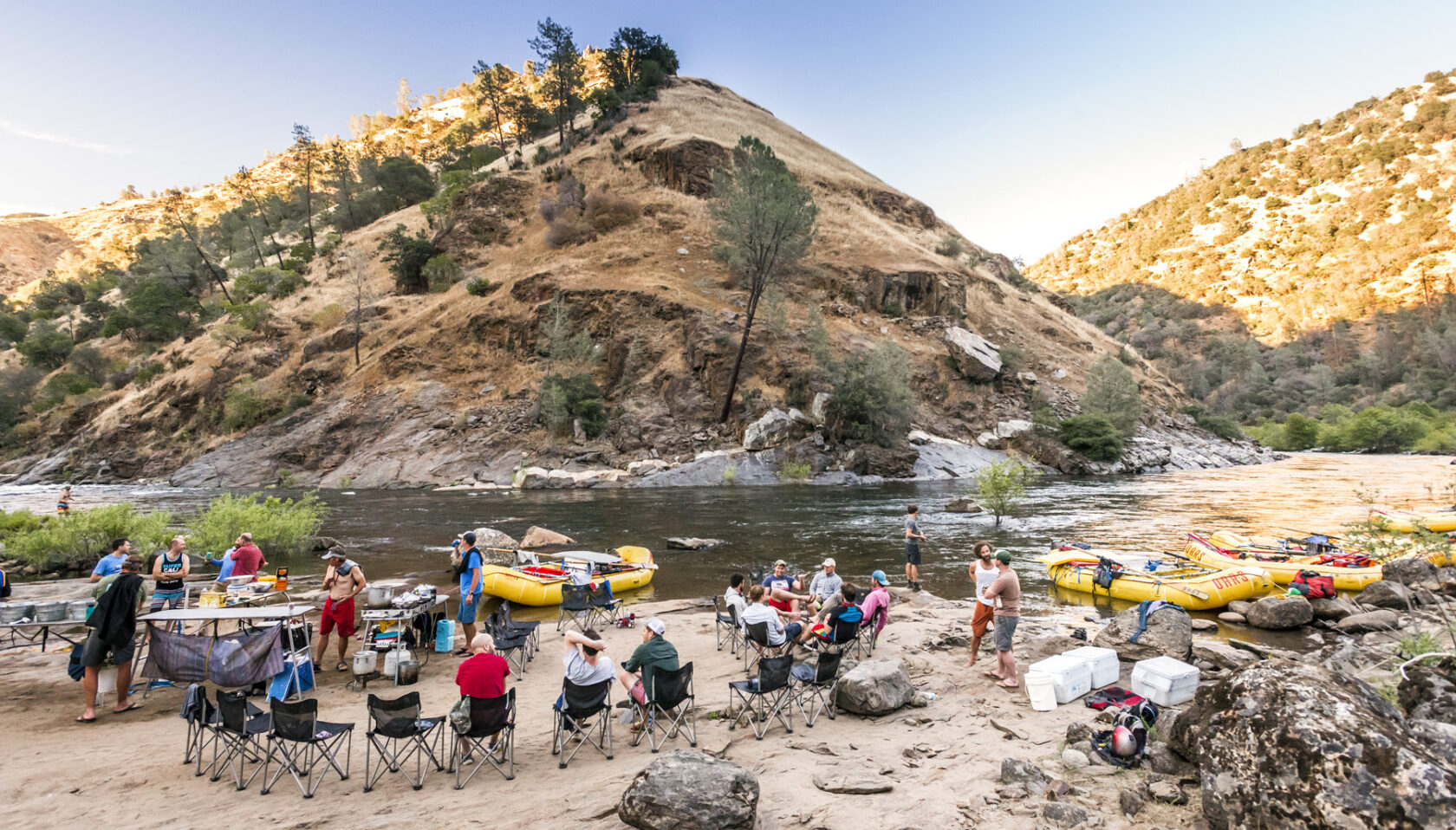 Group of people preparing a riverside meal with yellow rafts in the background, surrounded by hills and trees at sunset.