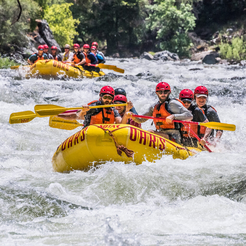 A group of people wearing helmets and life jackets paddle a yellow raft through wild rapids on the Tuolumne River near Yosemite