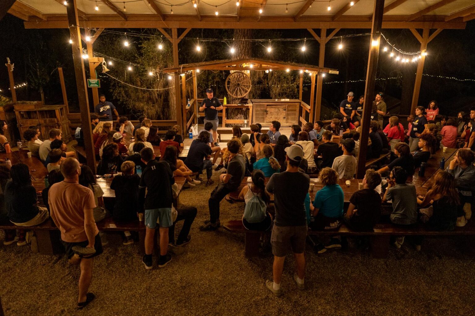 A large group of people sat around picnic tables listening to a man speaking on a small stage at night with string lights illuminating the scene.