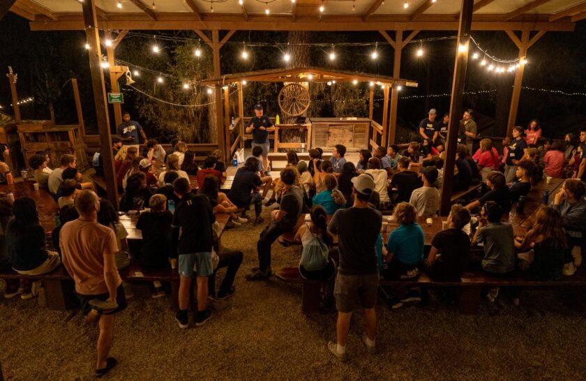 A large group of people sat around picnic tables listening to a man speaking on a small stage at night with string lights illuminating the scene.