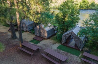 Canvas cabin tents lined up along the river amongst green trees.