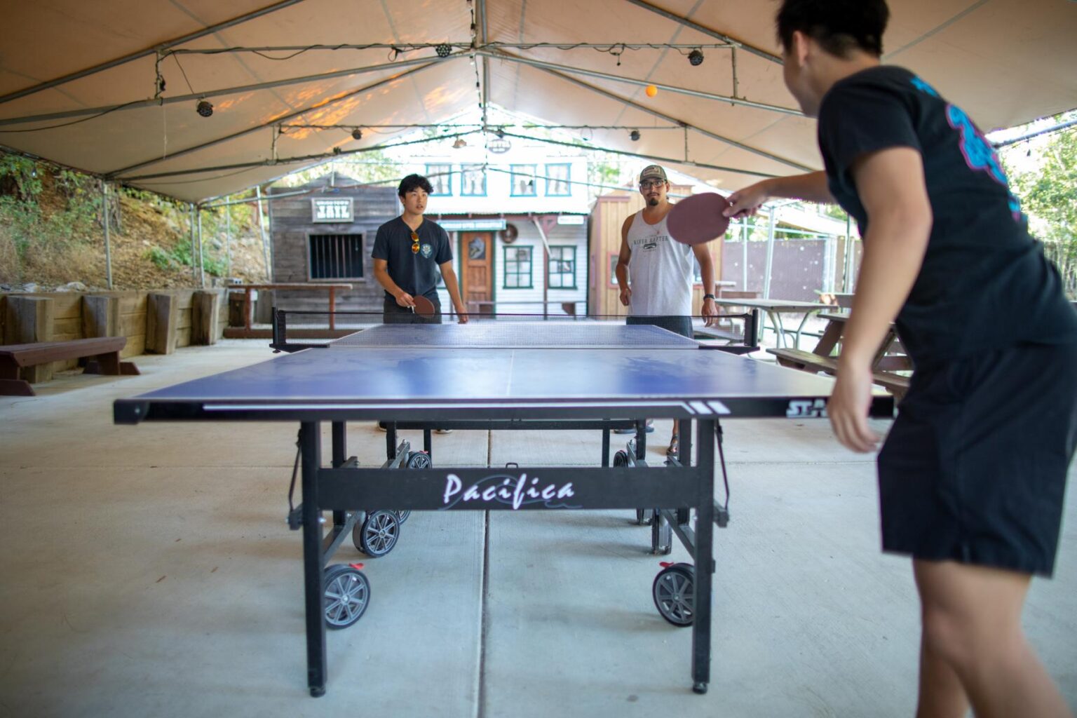Two young adults play pingpong at a table under a large canvas tent while an adult watches.