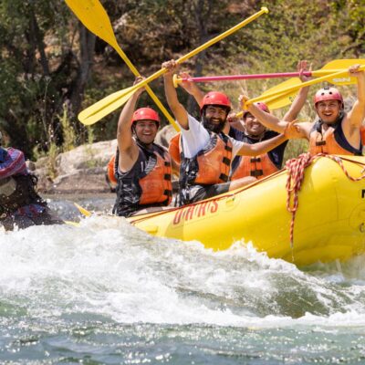 A group of rafters hold up their paddles while smiling coming out of a small rapid as the guide paddles.