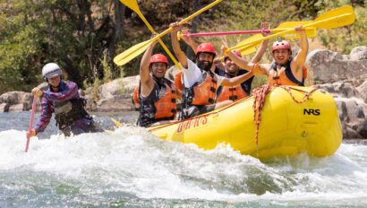 A group of rafters hold up their paddles while smiling coming out of a small rapid as the guide paddles.
