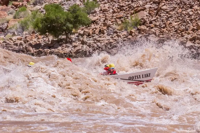 A white and red dory burst through a massive whitewater wave on the Colorado River as two passengers seated in the front hang on tight.