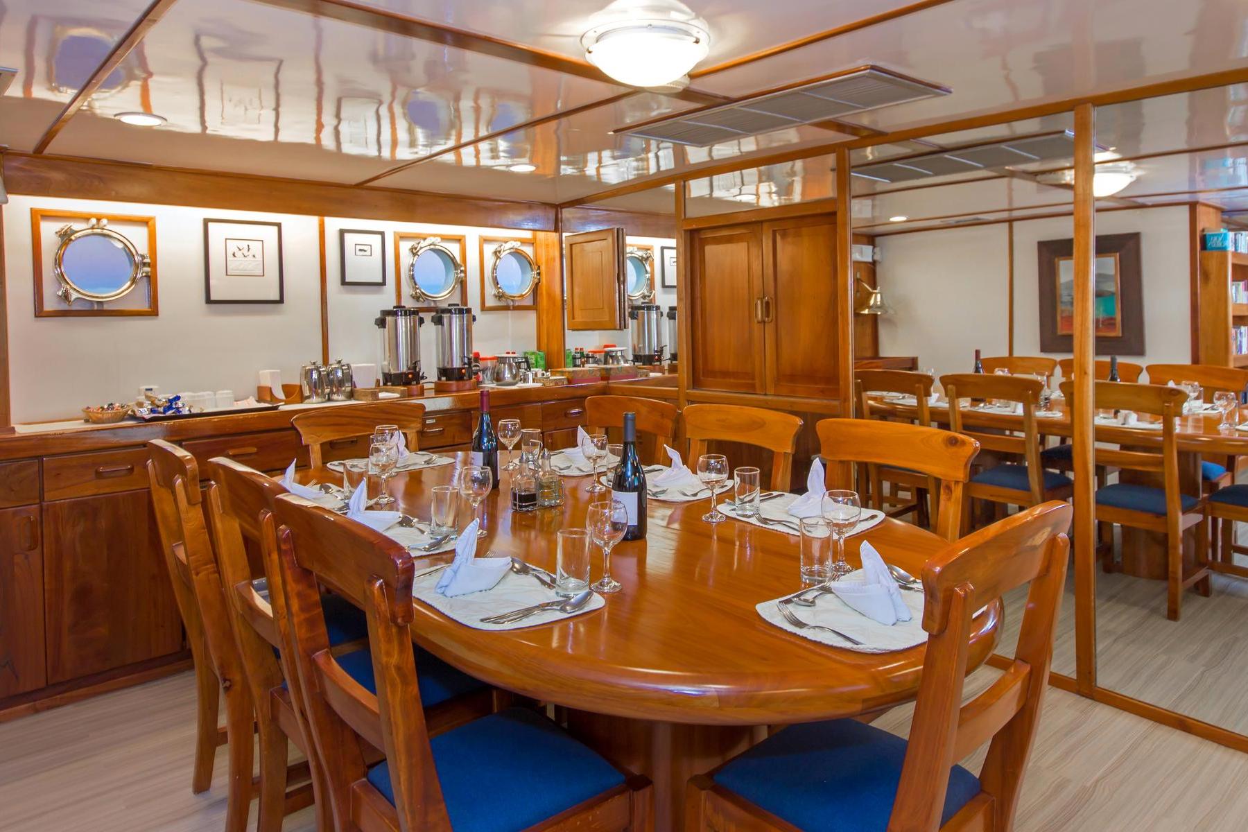 The dining room of the Beluga luxury yacht; 8 chairs suround a table with place settings, wine glasses, and bottles of wine. A beverage buffet runs along the wall behind it with 3 port holes looking over the ocean.