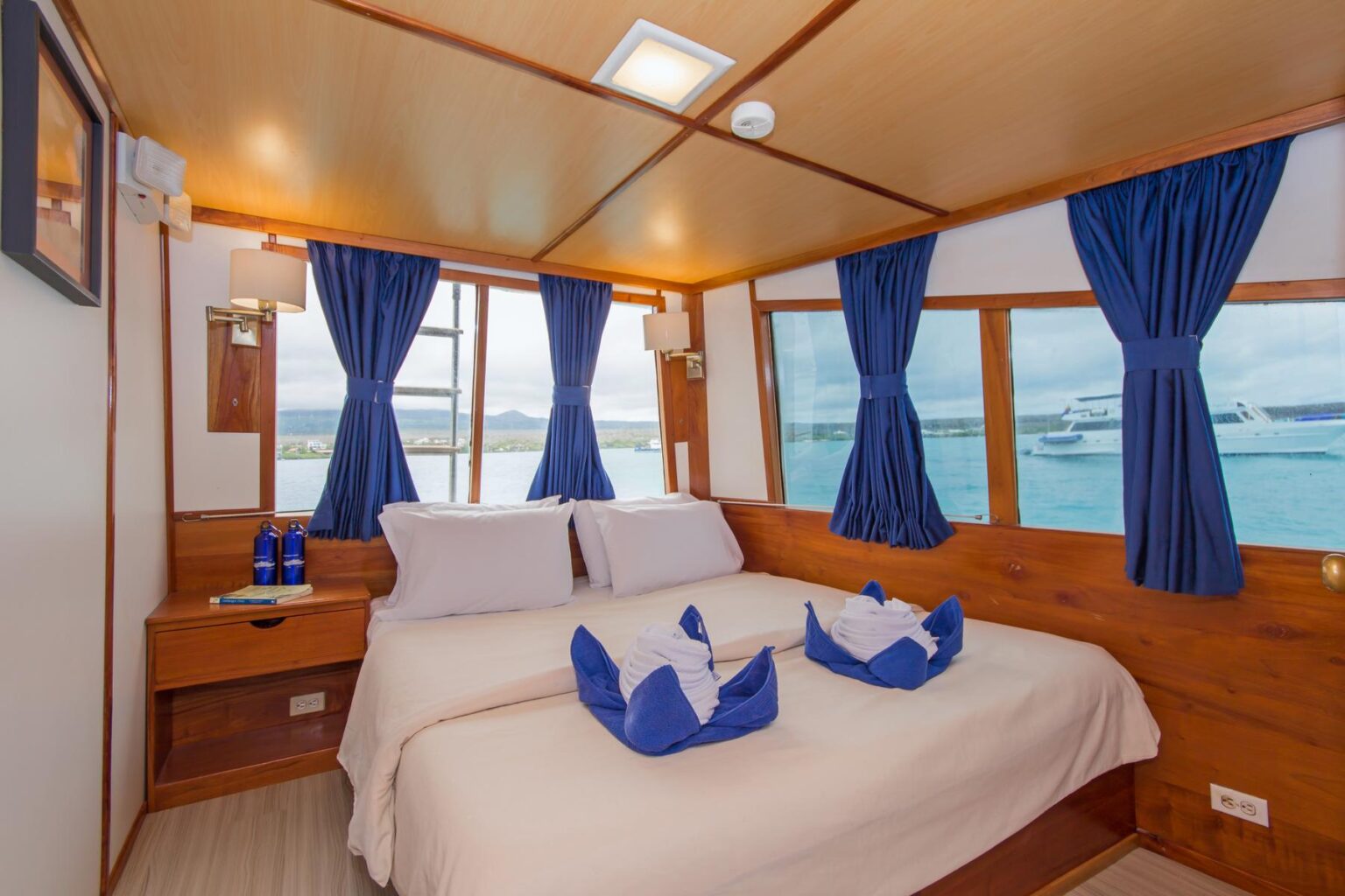 A room aboard the luxury yacht Beluga in the Galapagos islands; blue and white towels are decoratively displayed on the white comforter of a double bed and blue curtains are decoratively pulled back to reveal the ocean through large windows.