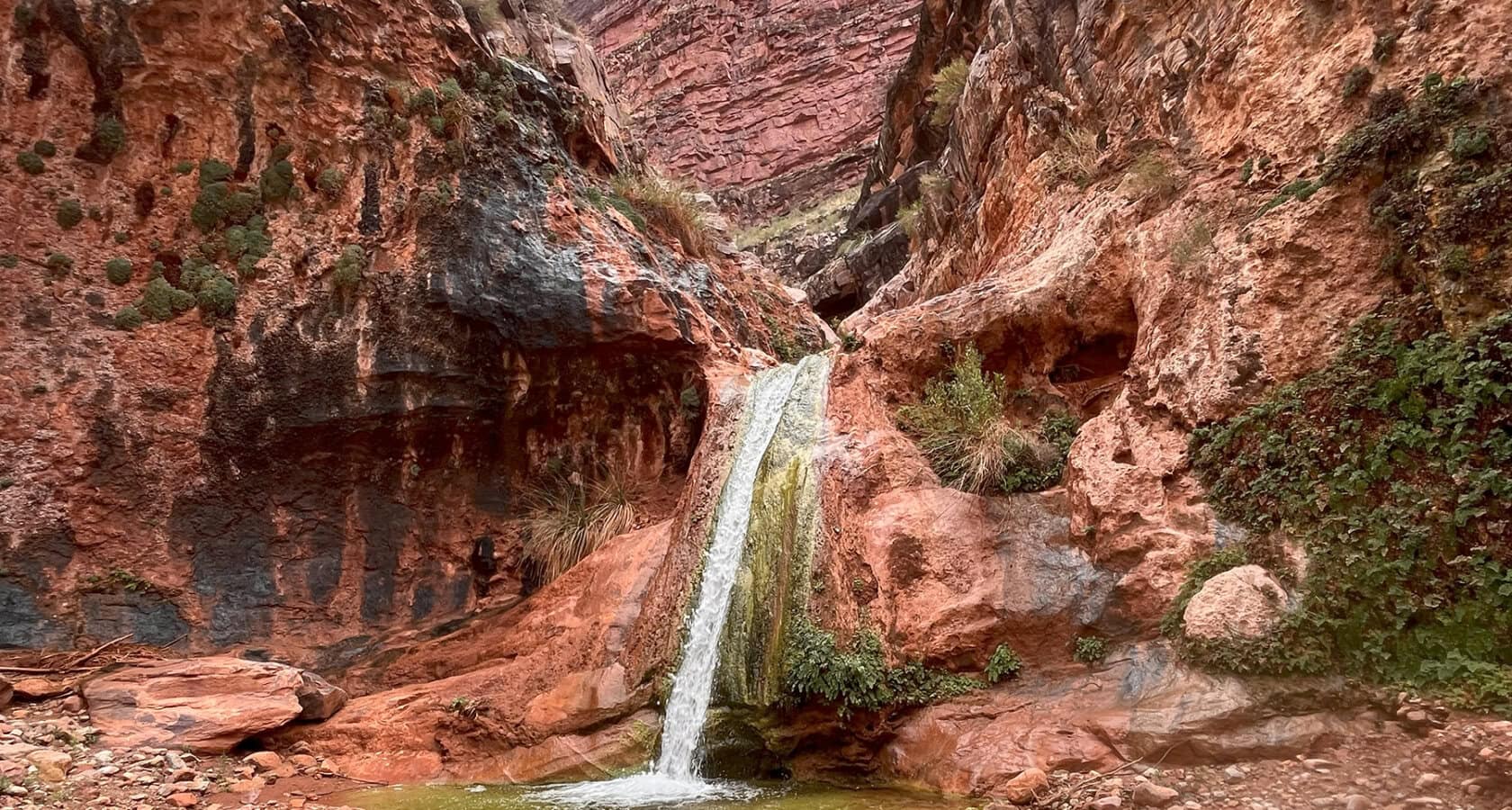 A waterfall spills over a ledge from a vibrant red rock canyon