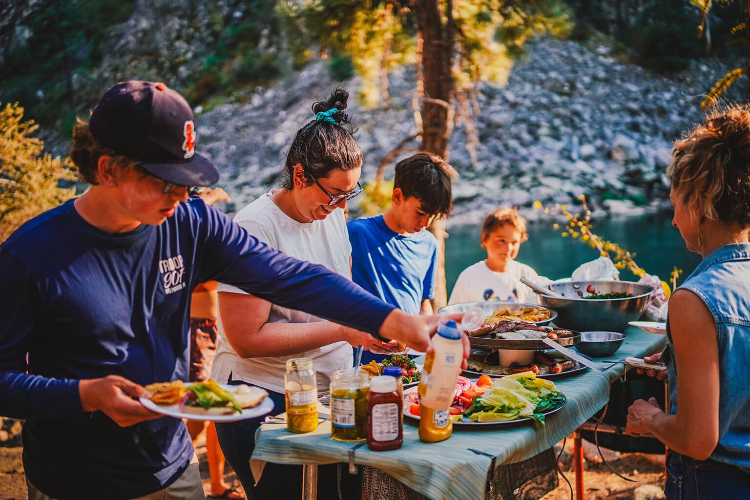 A family serves themselves hamburgers with all the fixings in a camp setting along a river