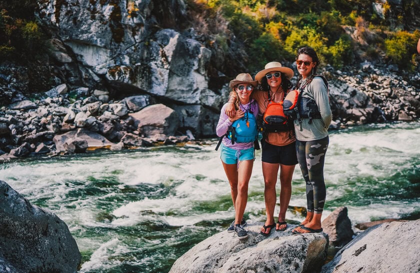 Three women in life jackets pose on a boulder next to a rushing river