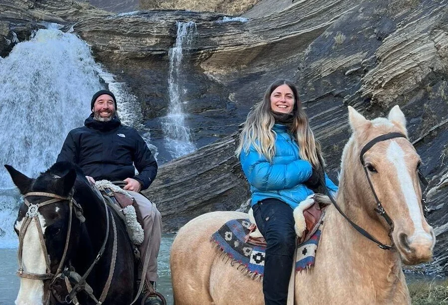 A man and a woman on horses smile at the camera in front of a waterfall in Patagonia