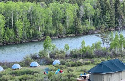 A serene campsite by a river, featuring tents, a large glamping tent, and people relaxing amidst lush greenery and trees.