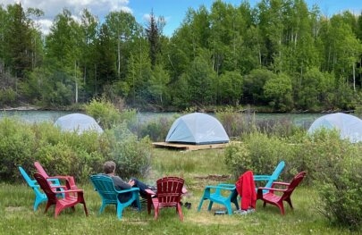 A man sits by a campfire overlooking a serene camp setting with white tents and a river flowing in the background