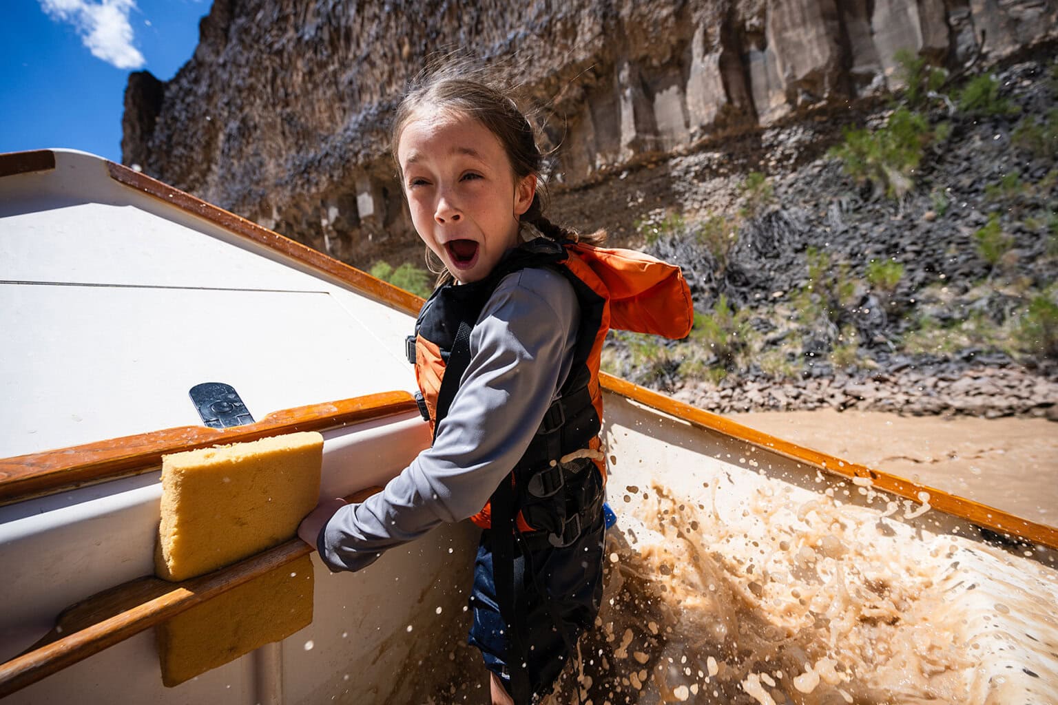 A little girl makes an excited face at the camera riding in a dory through whitewater