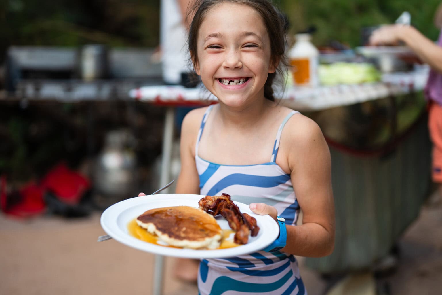 A young girl smiles at the camera as she holds up a plate of pancakes and bacon