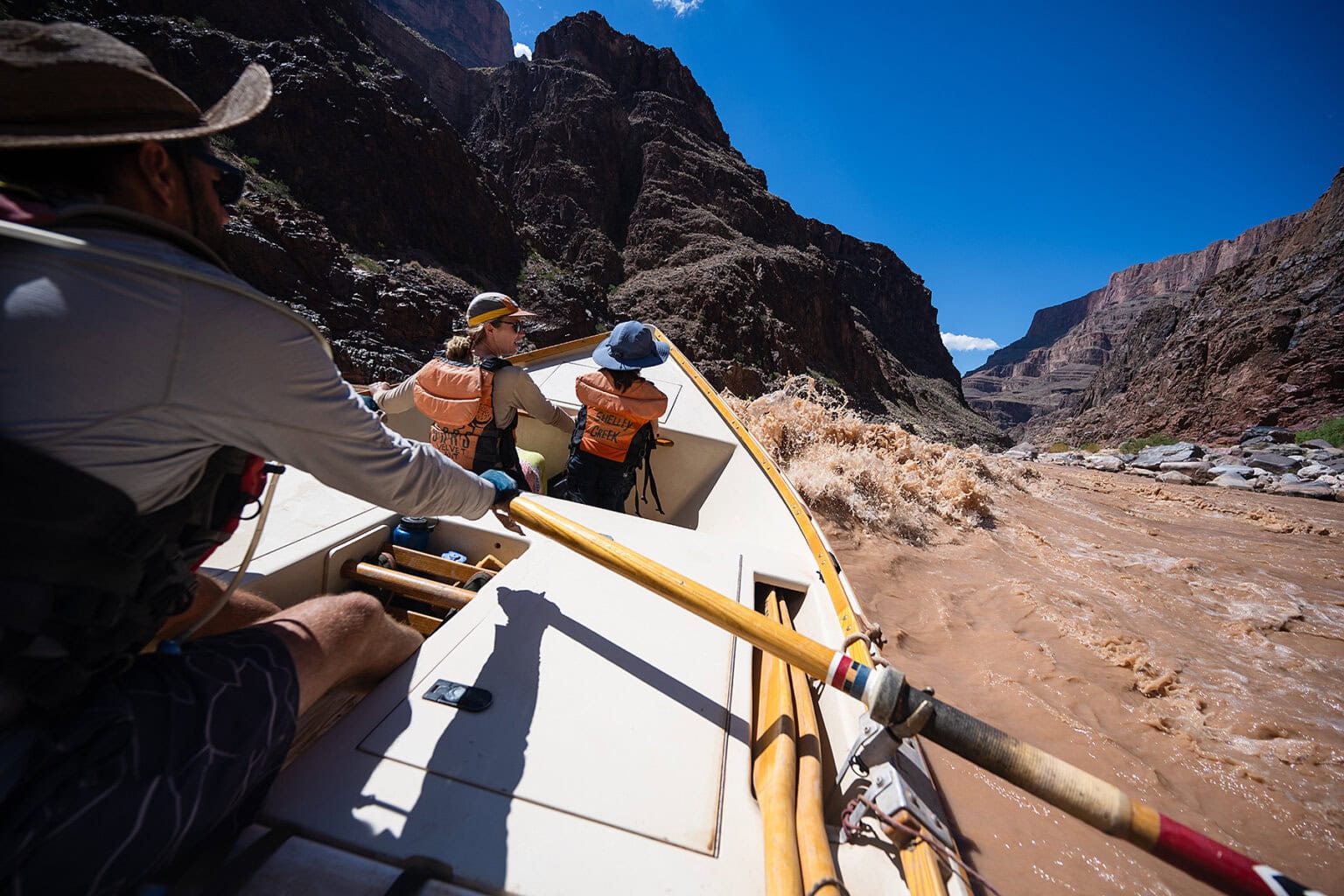 A man guides a dory with two passengers through a frothy rapid on a Grand Canyon river trip