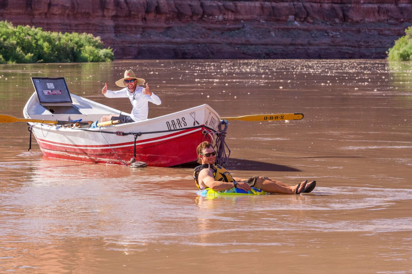 A man sits on a floaty in calm water on the Colorado River while a guide sits in a dory boat and gives thumbs up