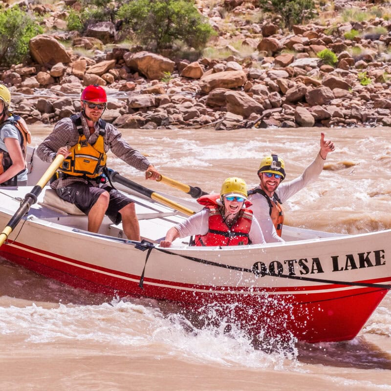 Three guests and a guide smile for the camera as they row through whitewater in a dory on the Colorado River through Cataract Canyon.