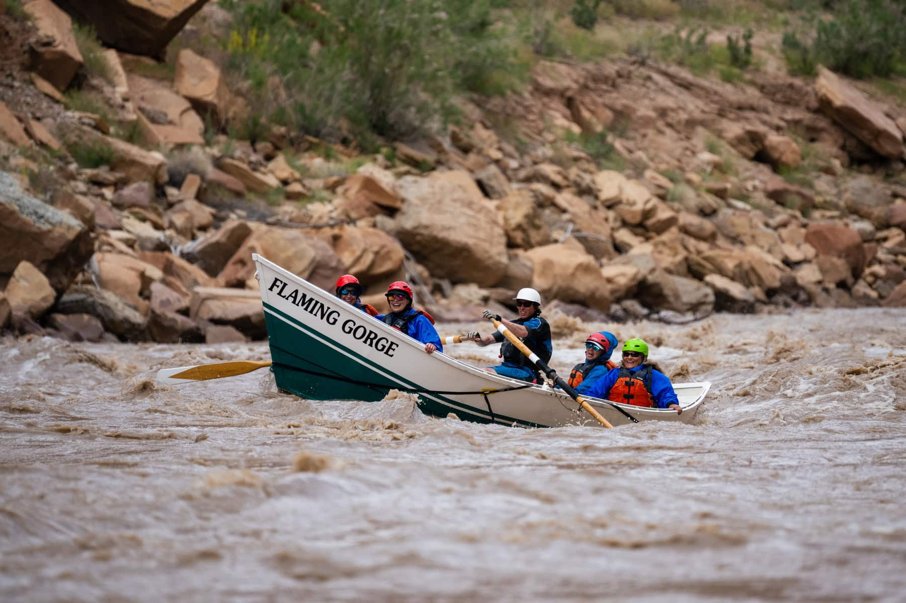 Four guests and a guide smile at the camera as they row through whitewater in a dory in Cataract Canyon.
