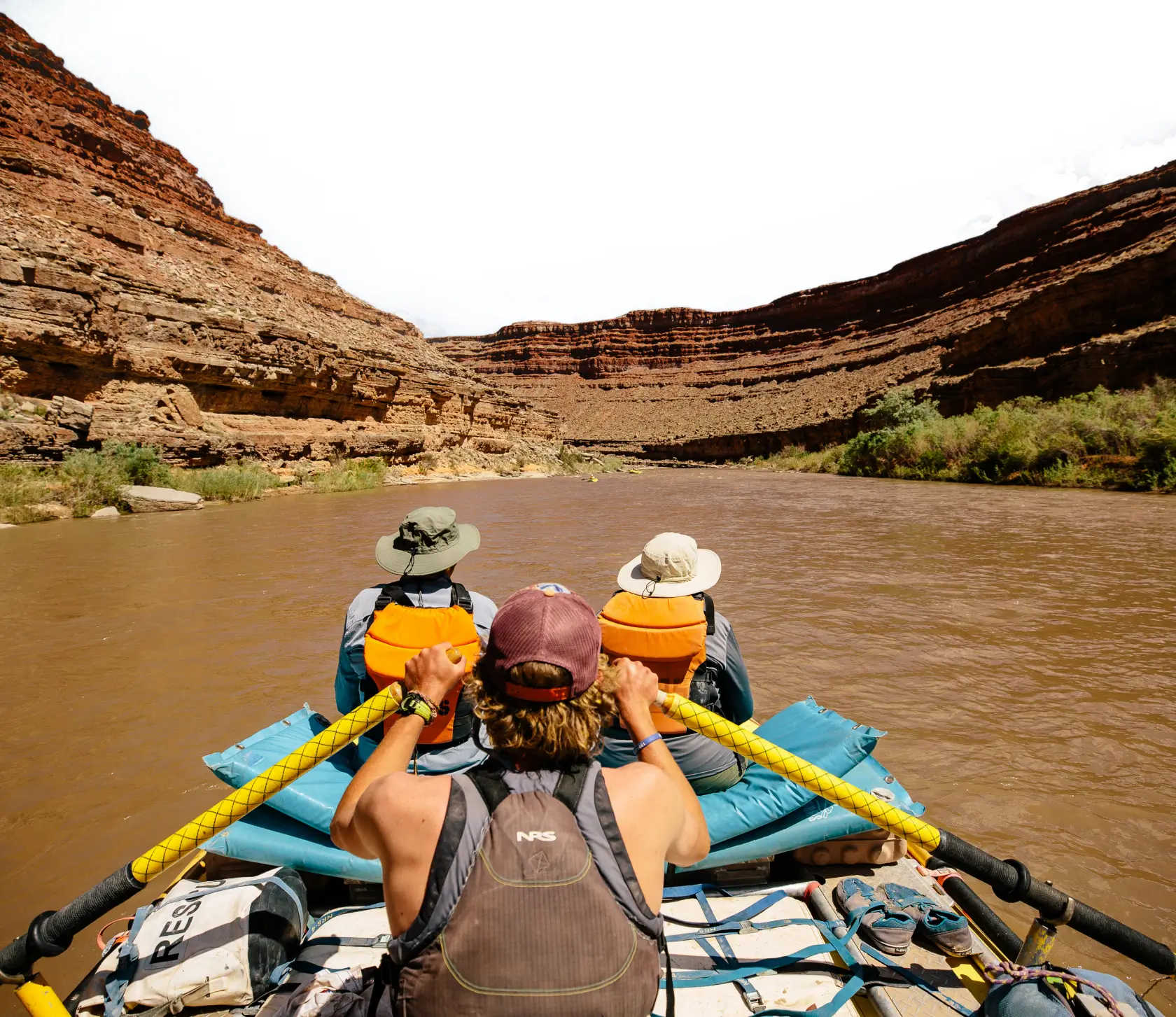 A group of rafters navigate the San Juan River surrounded by towering red rock cliffs under a bright sky.