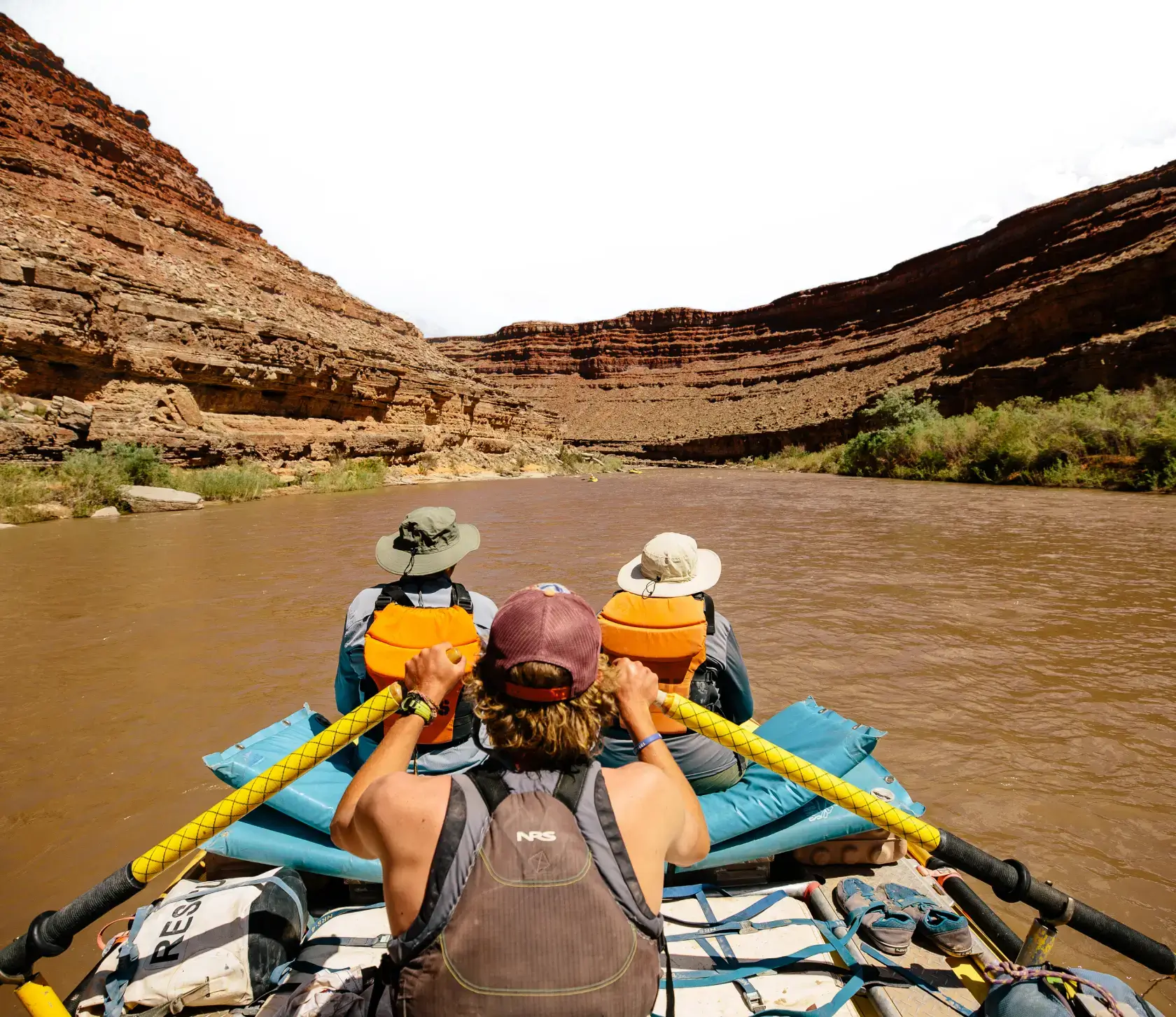 A group of rafters navigate the San Juan River surrounded by towering red rock cliffs under a bright sky.