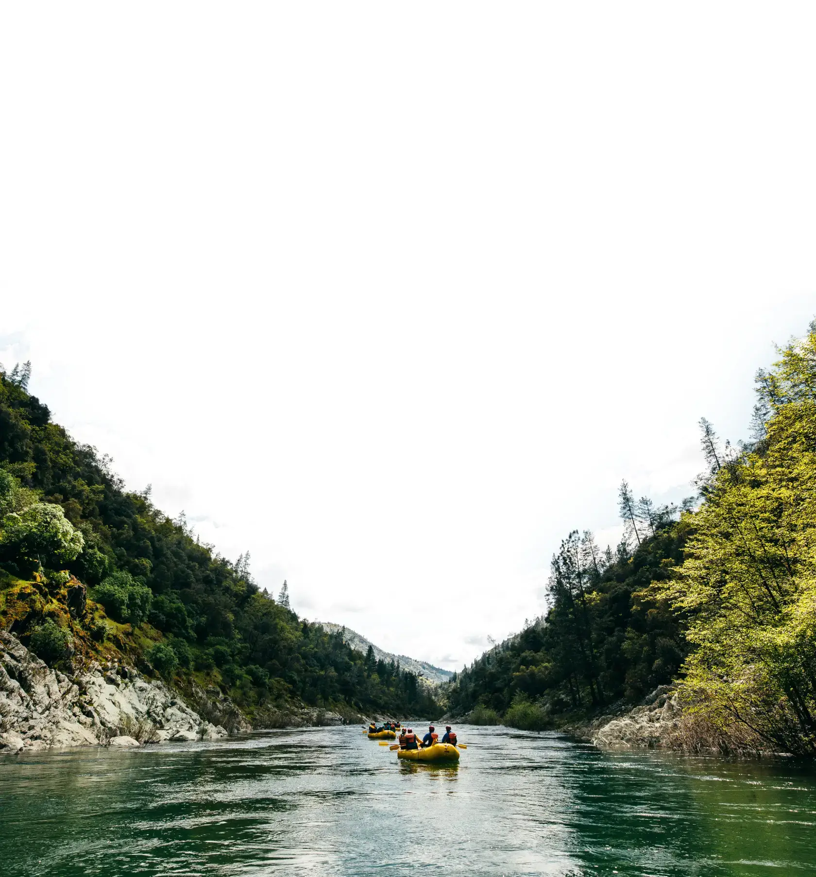A group of people in yellow rafts navigate a serene river surrounded by lush green hills and rocky banks under a cloudy sky.