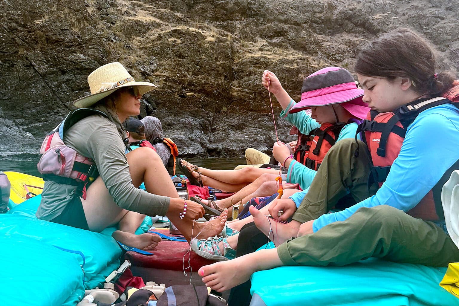 Several kids and a woman sitting on a raft making friendship bracelets. They are on a river and there's a rock wall in the background.