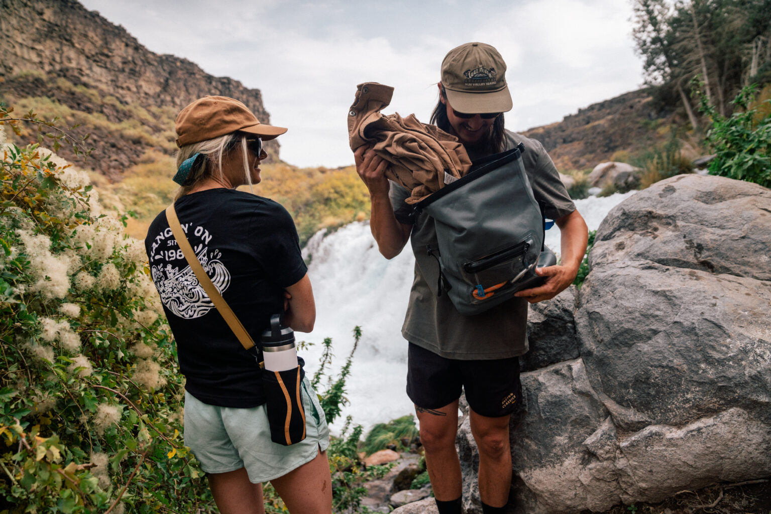 A hiker pulls gear out of a waterproof Chums Rolltop Sling bag while resting at the base of a waterfall with another hiker