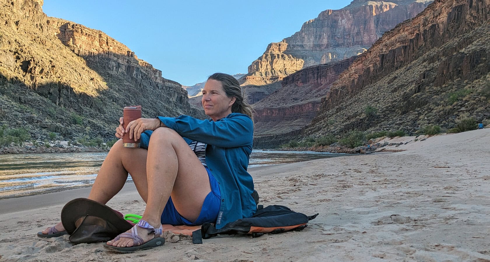 A woman sits on a sandy beach next to the Colorado River with towering cliffs rising behind her in Grand Canyon