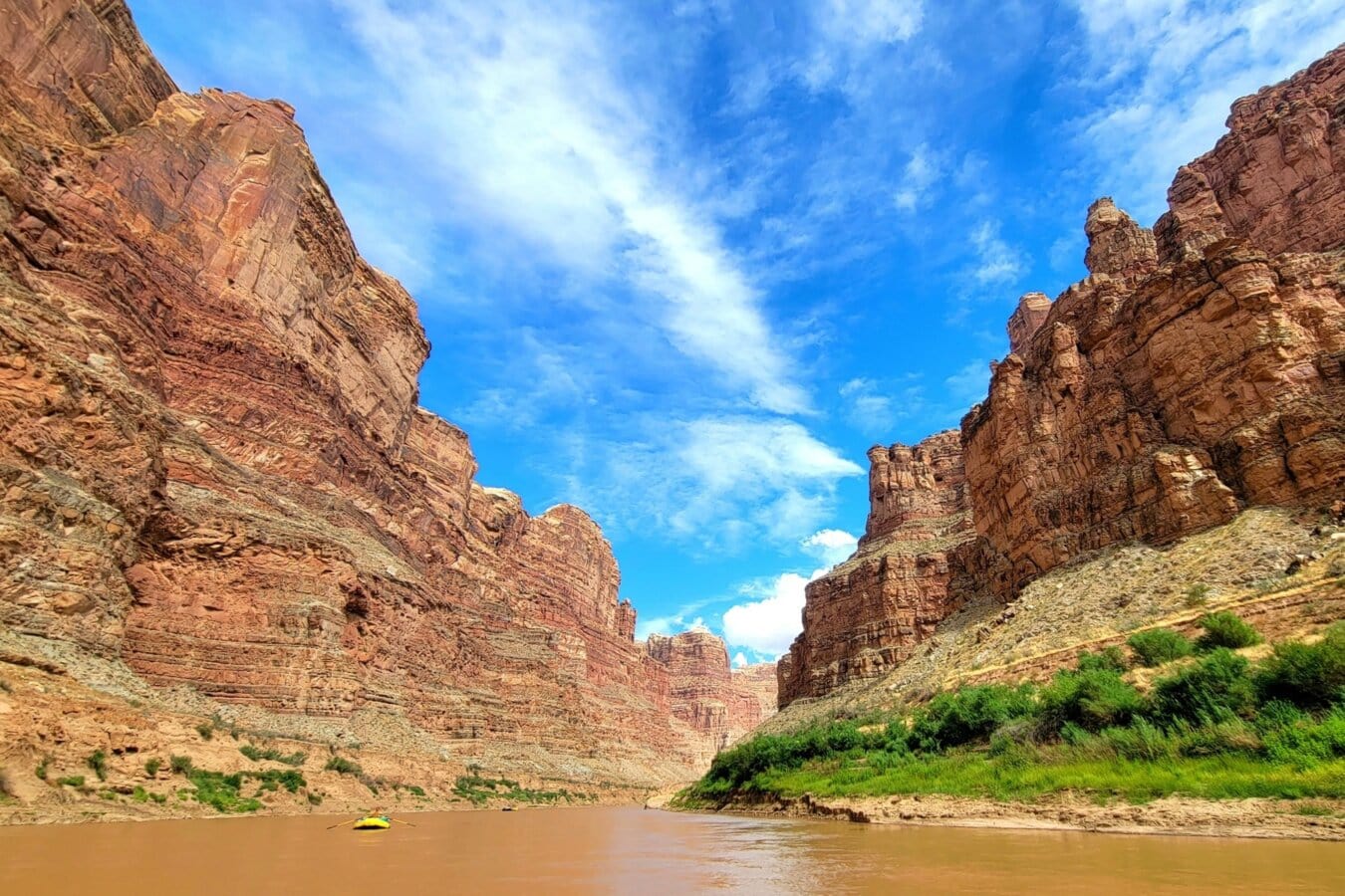A yellow raft rows down the Colorado River with towering canyon walls.