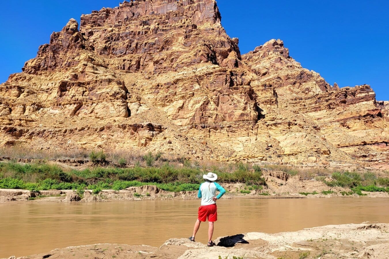A man stands on the banks of the Colorado River and looks up at the mountain across the river.