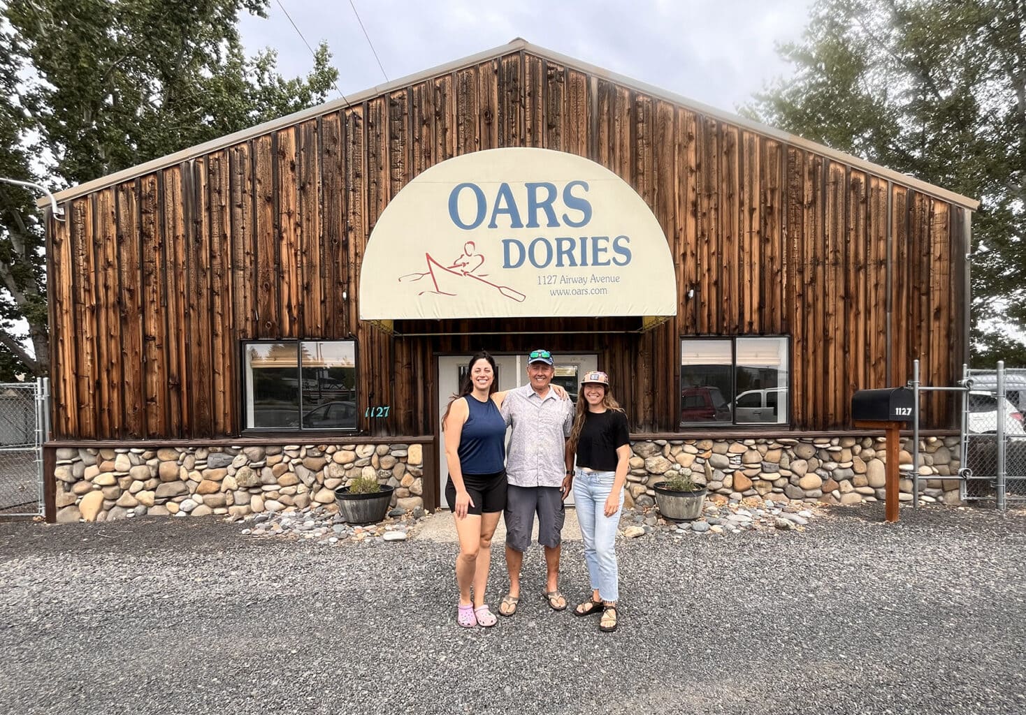 A man and two women pose in front of a wooden building with an OARS Dories Idaho awning hanging out front.