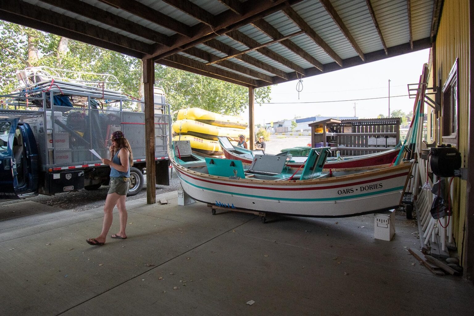 A woman standing in a comtemplative position in a wareshouse setting with dory boats and a truck filled with rafting gear in the background