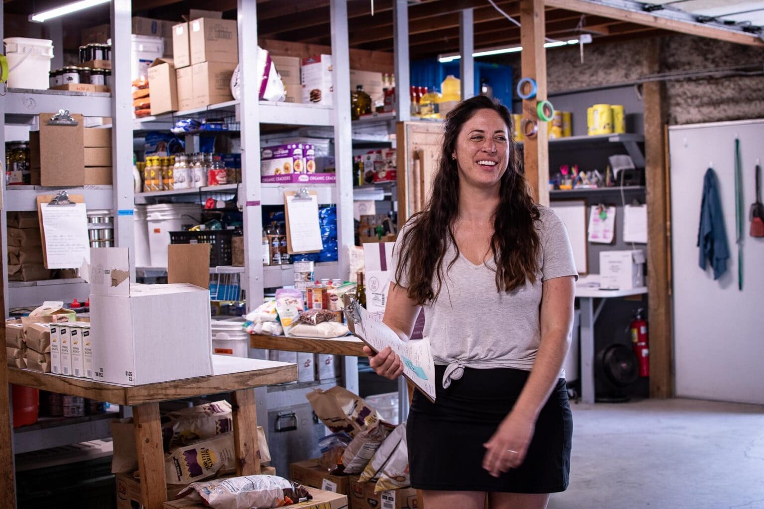 A woman stands in a rustic storage area stacked with food products.