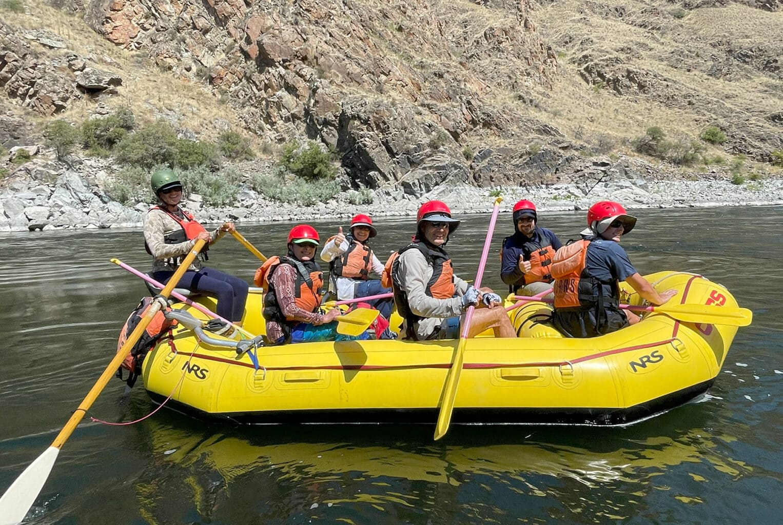 A group of people wearing helmets and life jackets are sitting in a yellow raft on a calm stretch of the Snake River in Idaho