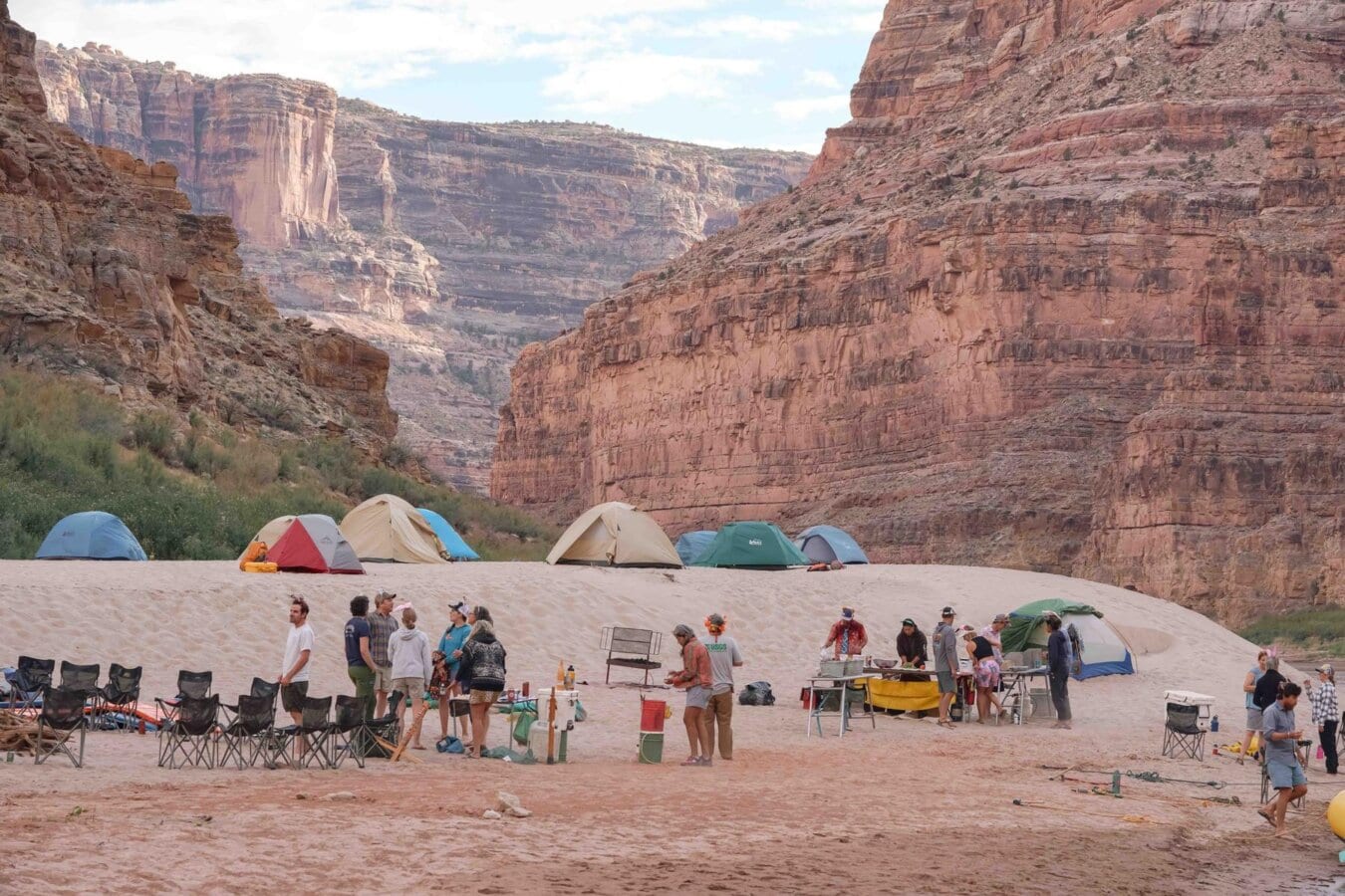 A group of campers hang out on a sandy beach in Cataract Canyon and several tents are set up on a sandy hill above them.