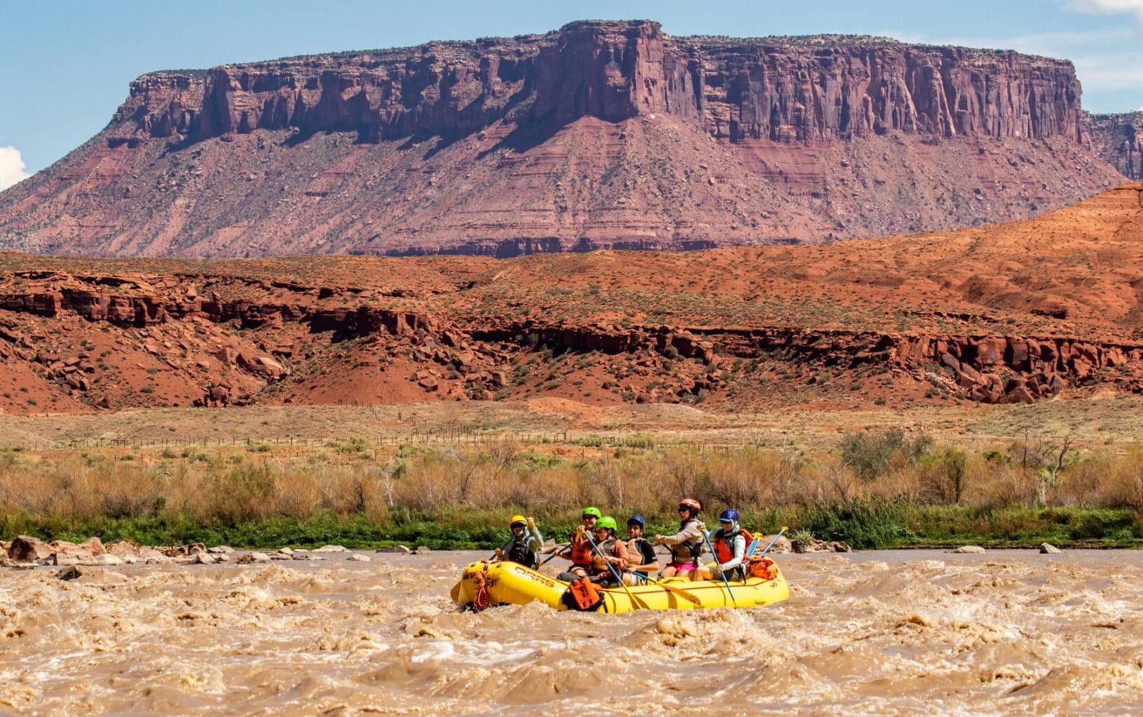 A group of paddlers in a yellow raft navigate small rapids on the Colorado River with a stunning desert canyon scenery in the background