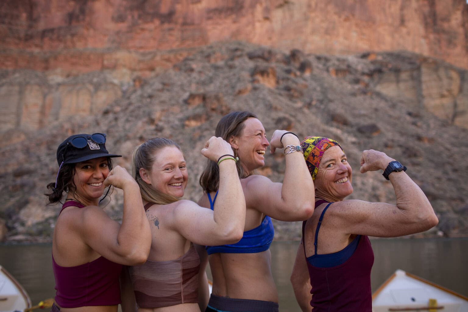 Four women Grand Canyon river guides flex their arms and smile in front of a rocky canyon and river, showing strength and camaraderie.