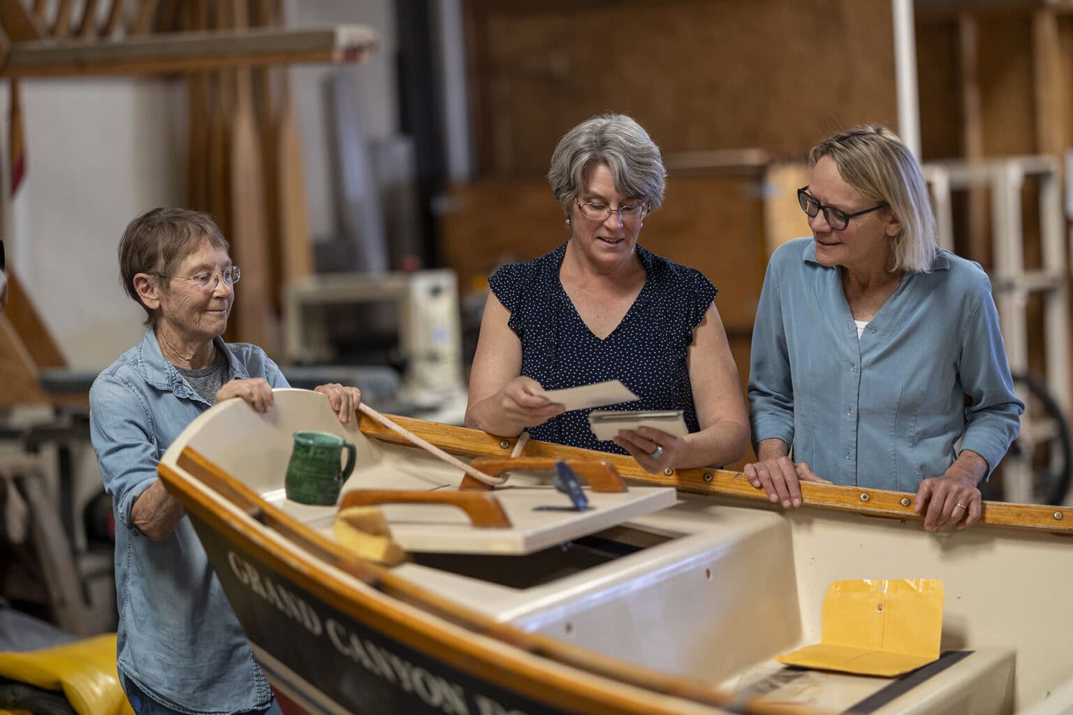Three women examine old photos while gathered around a wooden boat in a workshop.