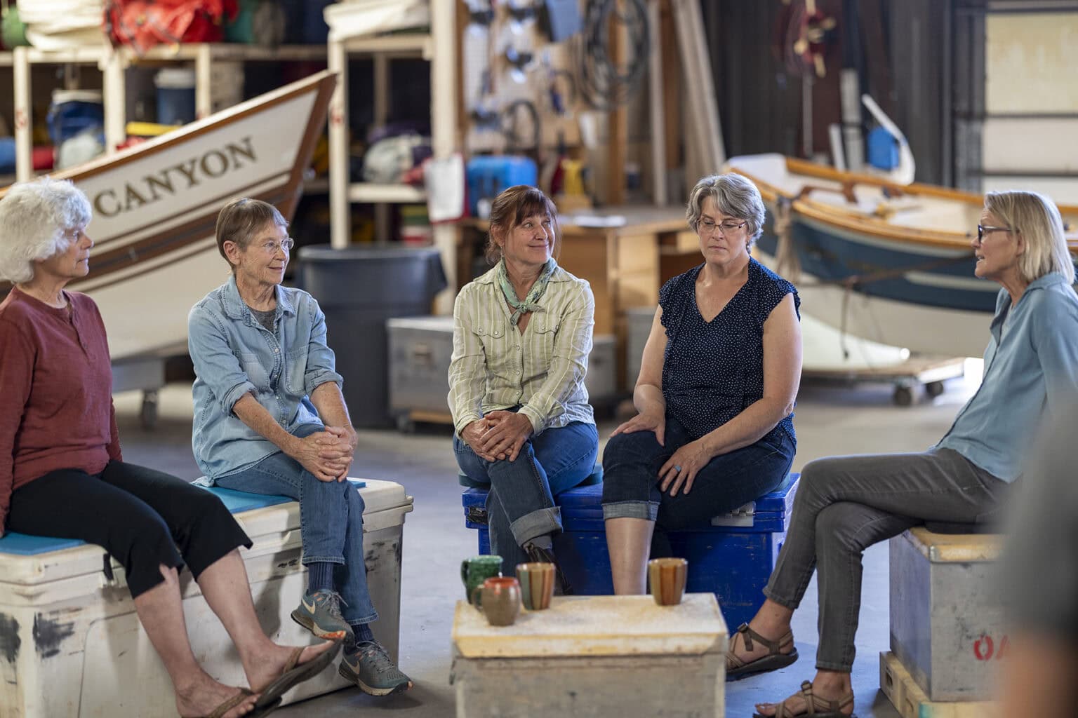 A group of five women seated on coolers and storage boxes gather in a warehouse storing dory boats.
