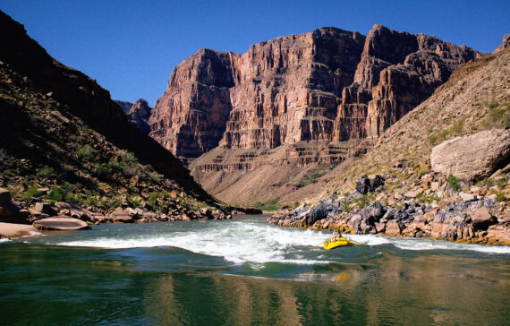 A river guide in a yellow raft navigates swirling waters in the Grand Canyon, surrounded by towering red rock cliffs under a clear blue sky.
