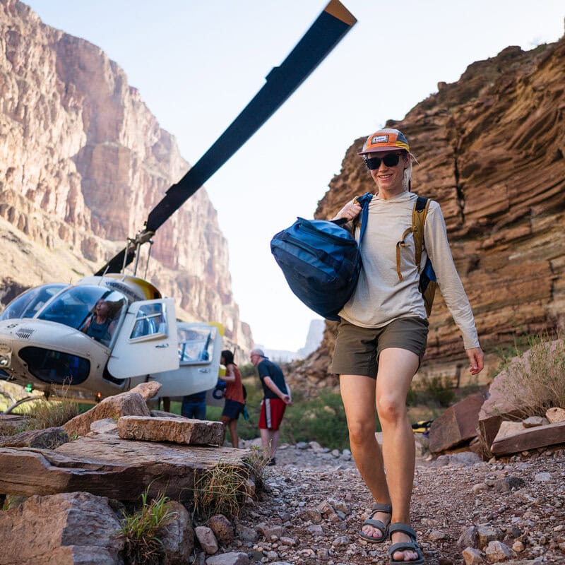 A person wearing shorts and a backpack walks towards a helicopter amidst towering canyon walls and rocky terrain in Grand Canyon.