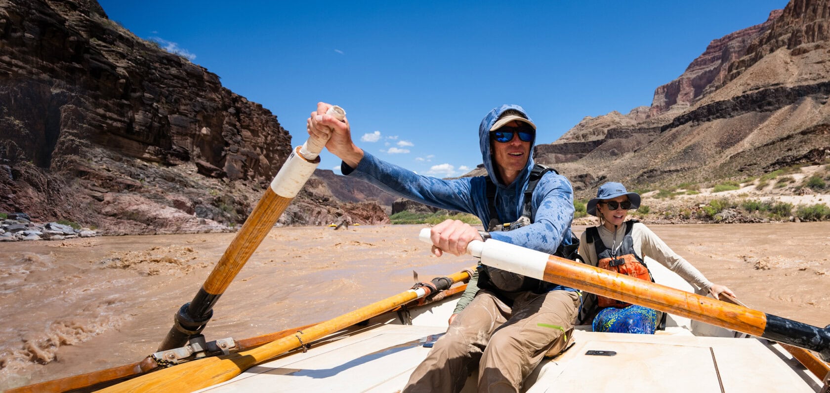 Two people in a wooden dory boat navigate the Colorado River surrounded by steep, rocky cliffs under a bright blue sky. Water splashes around them.