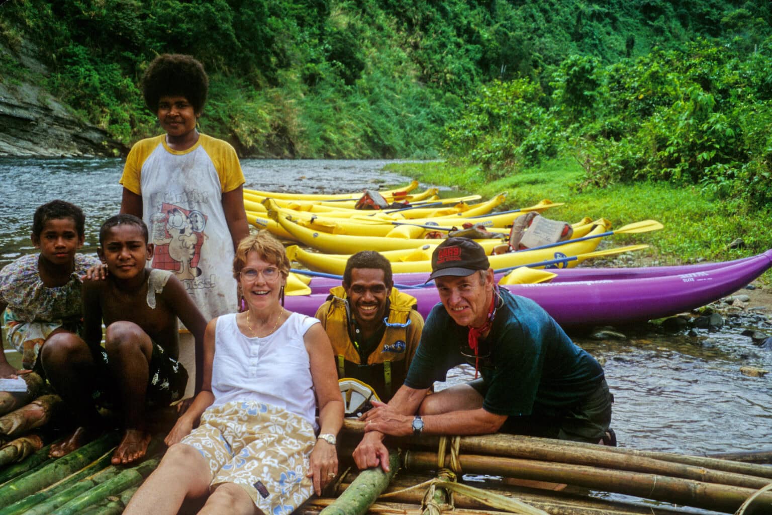 A group of six people, including locals and tourists, gathered on a bamboo raft near a river. In the background, there are bright yellow and purple kayaks along the riverbank, surrounded by dense green vegetation. 