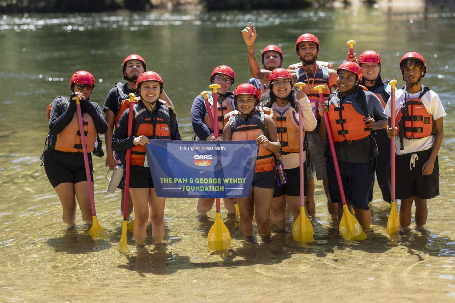 A group of high school students stands in shallow water, wearing life jackets and helmets, holding paddles and a banner for the Pam & George Wendt Foundation.