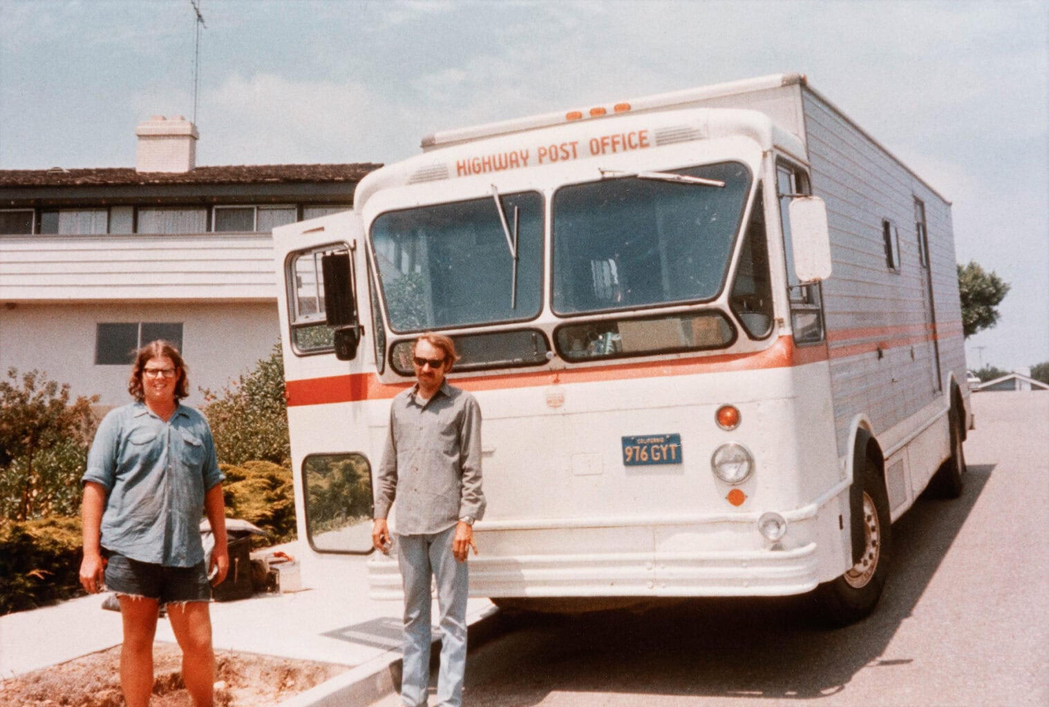 Two men stand in front of a vintage Highway Post Office truck on a neighborhood street