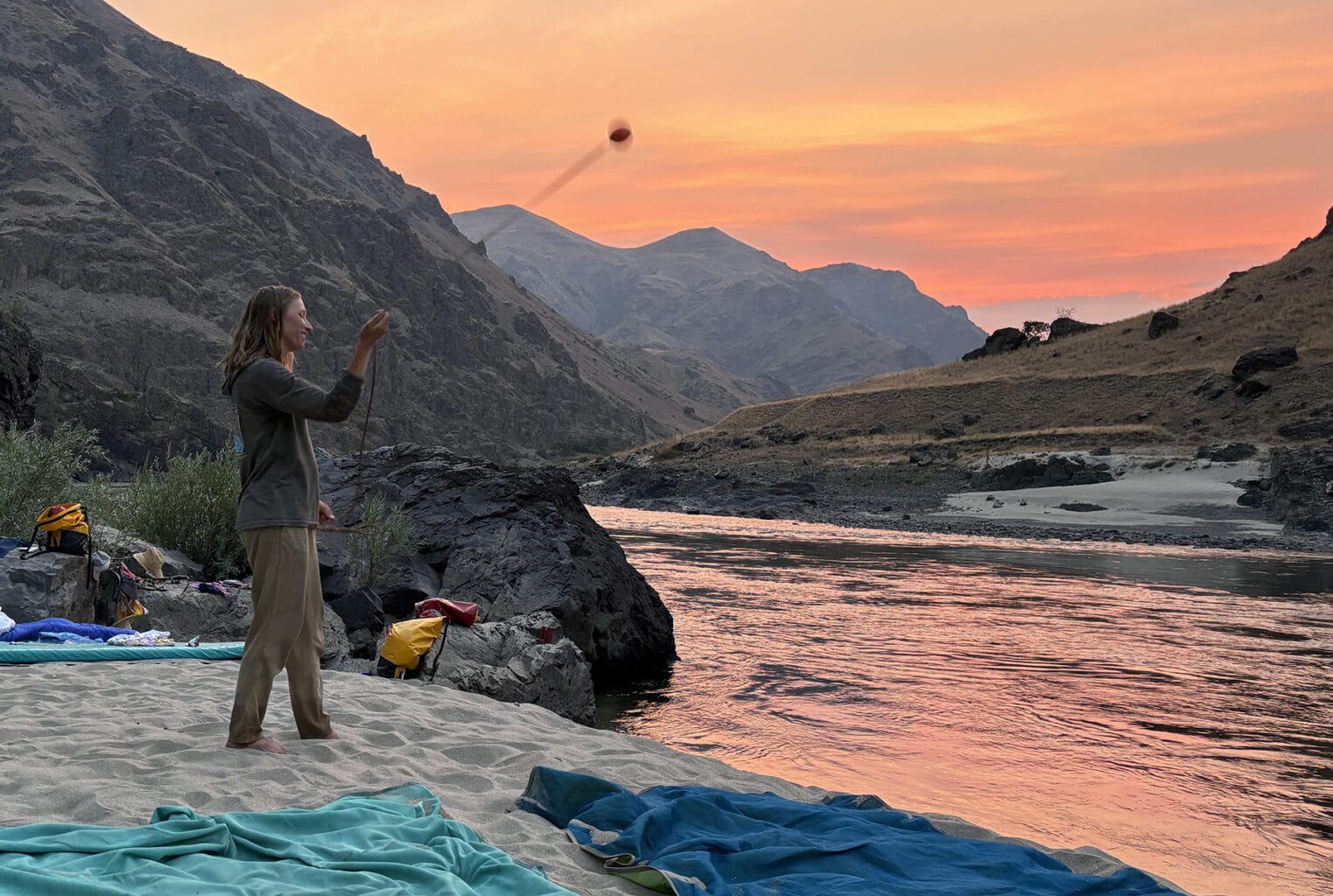A woman gives a dart ball performance on a sandy beach alongside a river at sunset.