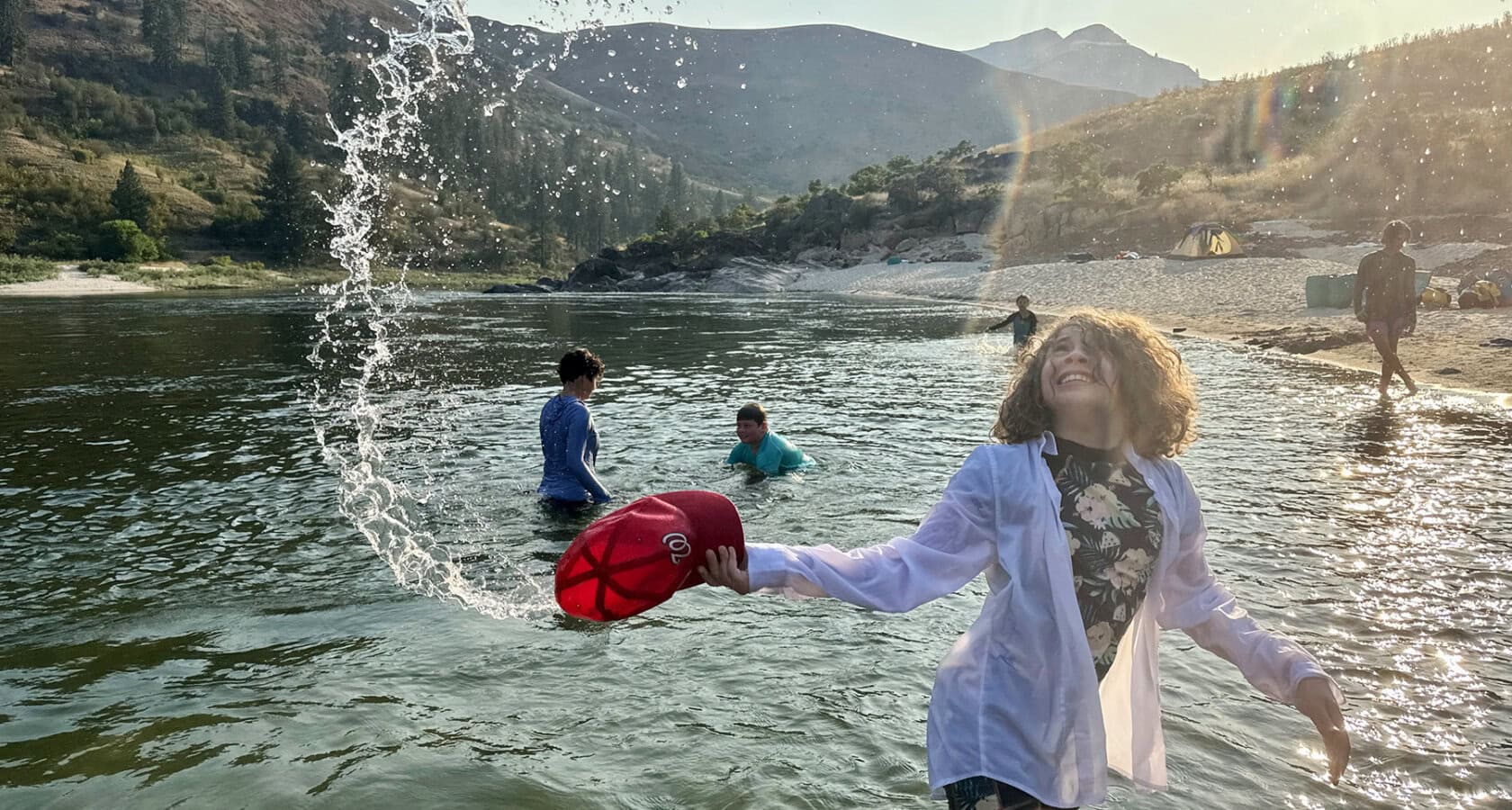 A girl splashes on the shore of a river with people frolicking in the shallow water in the background